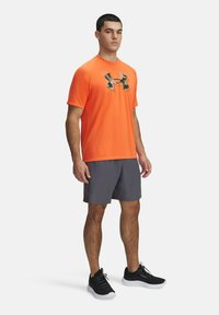 Orangefarbendes Sport-T-Shirt mit einem Camouflage-Logo, kombiniert mit grauen Shorts. Das Modell trägt schwarze Sneaker mit weißen Sohlen. Sauberes, modernes Design.
