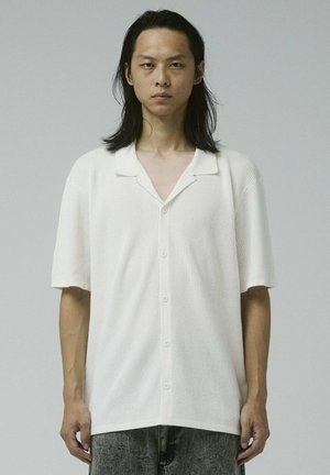 Uomo con lunghi capelli neri, indossa una camicia bianca a maniche corte con bottoni e pantaloni grigi, in piedi davanti a uno sfondo grigio uniforme.
