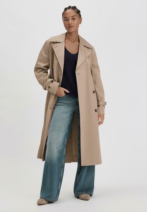 Beiger Trenchcoat mit breiten Revers und doppelter Knopfleiste, kombiniert mit einem dunkelblauen, gerippten Top und weit auslaufenden blauen Jeans mit leichten Used-Effekten.