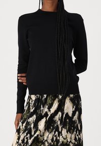 Zwarte gebreide trui met lange mouwen en een ronde halslijn, gecombineerd met een patroonrok in tinten groen, zwart en crème.