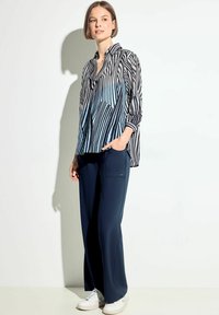 Blusa a strisce bianco e nero con dettagli blu, maniche lunghe e colletto, abbinata a pantaloni a gamba larga blu navy e sneakers bianche.