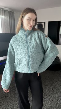 Fleece jacket in light blue with a textured pattern, high collar, and full zipper; features oversized sleeves and subtle branding on the shoulder.