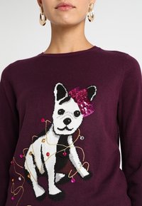 Violet neulepaita, jossa on musta-valkoinen koira-kuvio, vaaleanpunaisella helmillä varustettu rusetti sekä värikkäitä koristehelmiä ja -nyörejä.