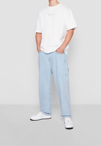 Man som bär lösa ljusblå jeans, vit t-shirt med grafik, vita sneakers, står med händerna i fickorna framför en enfärgad bakgrund.