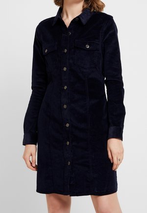 Vrouw draagt een marineblauwe corduroy jurk met knopen, lange mouwen en twee klepzakken op de borst.