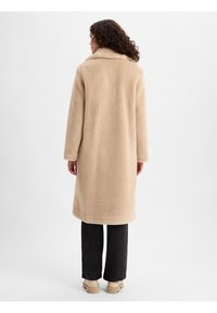 Beżowy płaszcz z futra syntetycznego z kołnierzem, długimi rękawami i prostym krojem. Materiał jest miękki, o długości do kolan i bocznych kieszeniach.