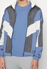 Młoda osoba w niebieskiej koszulce i czarnej, niebieskiej oraz białej kurtce zapinanej na zamek z elastycznymi mankietami i jasnoszarych spodniach.