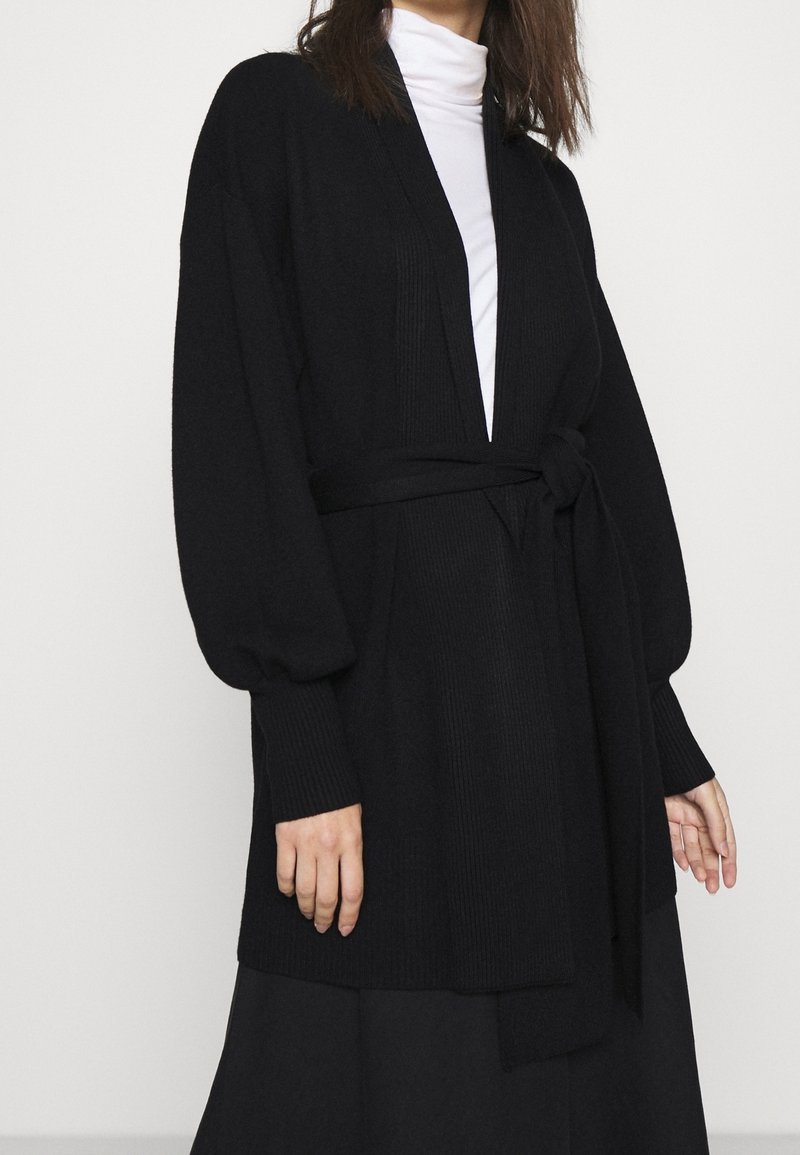 Cardigan noir tricoté avec de longues manches bouffantes, un décolleté en V profond et une ceinture nouée à la taille. Tissu texturé avec une finition lisse.