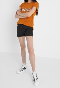 Oranžové tričko s bílým grafickým textem, černé šortky a bílé vysoké tenisky s černým nápisem. Jednoduchý, ležérní outfit se moderním vzhledem.