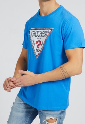 Homme portant un t-shirt bleu vif avec un grand logo blanc triangulaire GUESS et un jean bleu clair délavé, mains jointes devant.