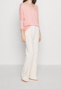 Pull en V rose clair avec des manches 3/4 associé à un pantalon large crème. Le pull présente une coupe décontractée et une texture douce. Chaussures accents orange.