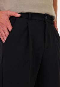 Zwarte geplisseerde broek met een gladde textuur, voorzien van een riemlus, knoopsluiting en zijzakken, gecombineerd met een beige top.