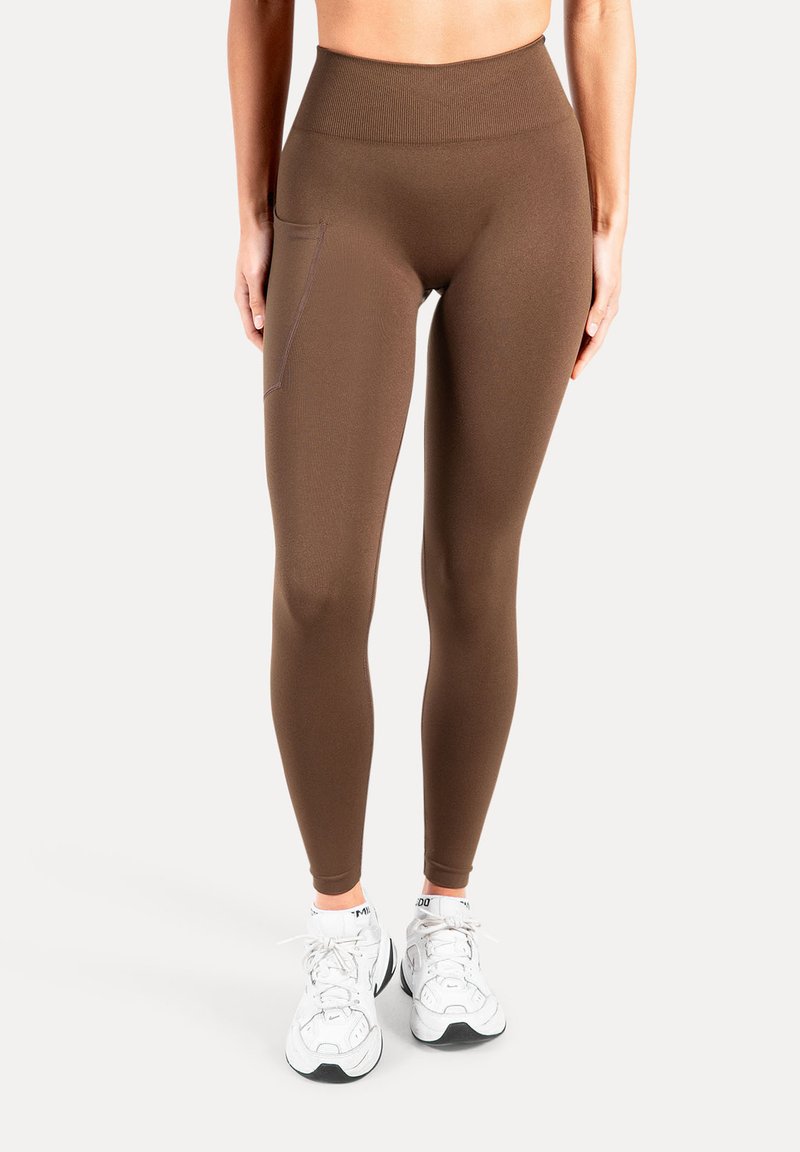 Bruine leggings met hoge taille gemaakt van rekbare, gladde stof, voorzien van een zijzak en een taps toelopend model. Draagt witte sportschoenen.