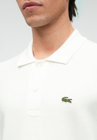 Polo shirt blanc en tissu texturé, avec un col à trois boutons et un petit logo de crocodile vert sur la poitrine gauche.