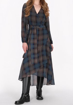 Frau trägt ein langärmliges, blau-rot kariertes Wickelkleid mit asymmetrischem Saum, kombiniert mit schwarzen kniehohen Stiefeln mit Schnallen.