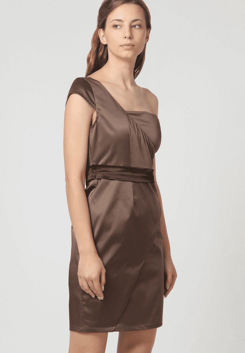 Mujer vestida con un vestido de satén marrón sin mangas y de un solo hombro, con una cintura ajustada, mirando ligeramente hacia su derecha sobre un fondo liso.