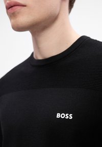 Detailný záber osoby v čiernej ribbovej svetri s bielym logom "BOSS" na hrudi, zobrazuje líniu čeľuste a krk.