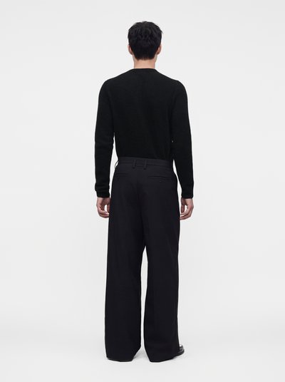 Man wearing a black long-sleeve sweater and wide-leg black trousers stands facing away on a plain white background.