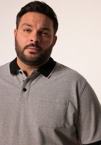 Harmaa polo-paita mustalla kauluksella, jossa on tasku vasemmalla puolella, teksturoitu kangas ja kolmen napin lista.