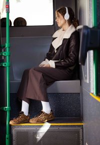 Hnědý vlněný kabát s krémovým límcem z beránčí kožešiny, doplněný dlouhou hnědou sukní, bílými ponožkami a teniskami s leopardím vzorem; viditelný zelený autobusový sloupek.