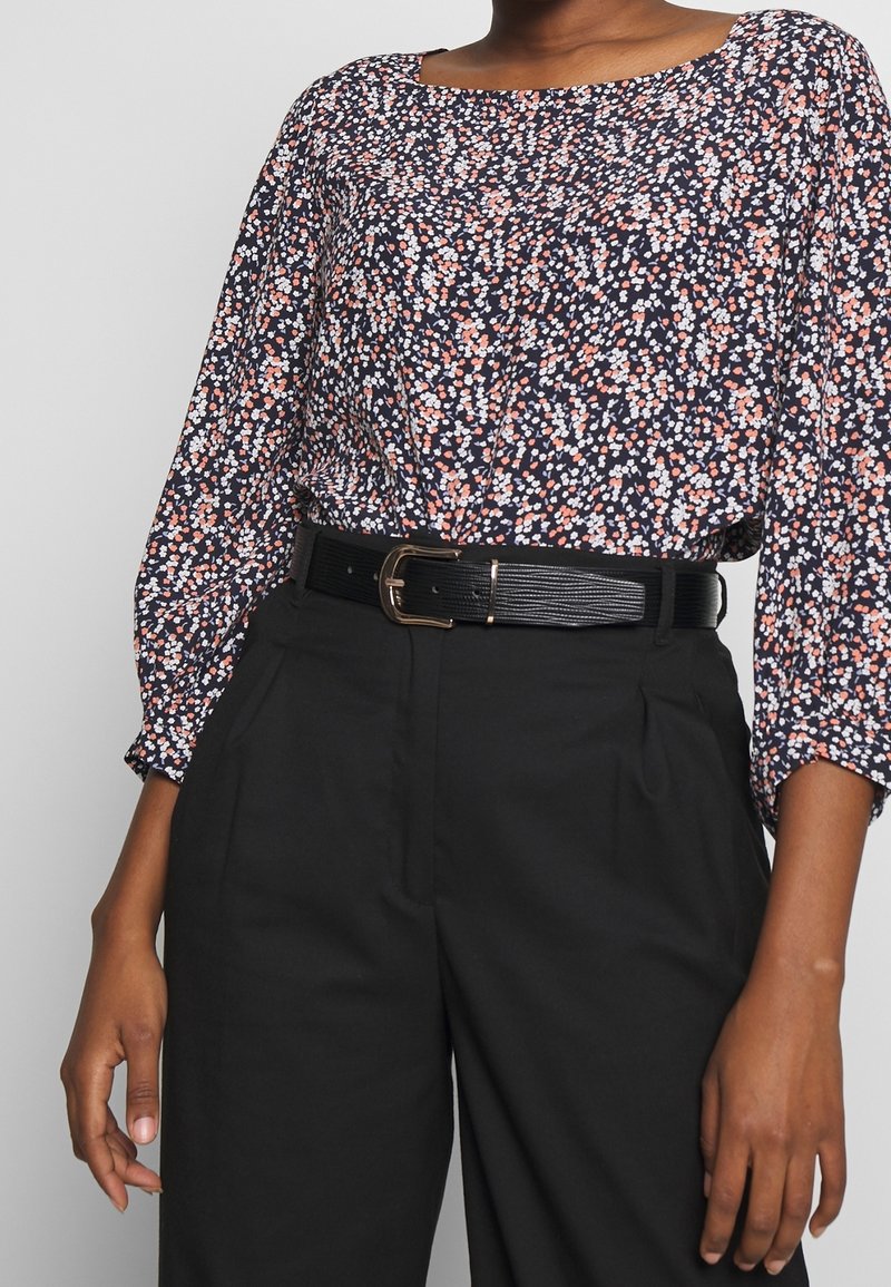 Femme portant un chemisier à fleurs noir, blanc et rose rentré dans un pantalon noir taille haute avec une ceinture en cuir noire.