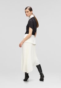 Black draped blouse with white collar, paired with a white pleated skirt. Model wears knee-high black heeled boots. Textured fabric details visible.
