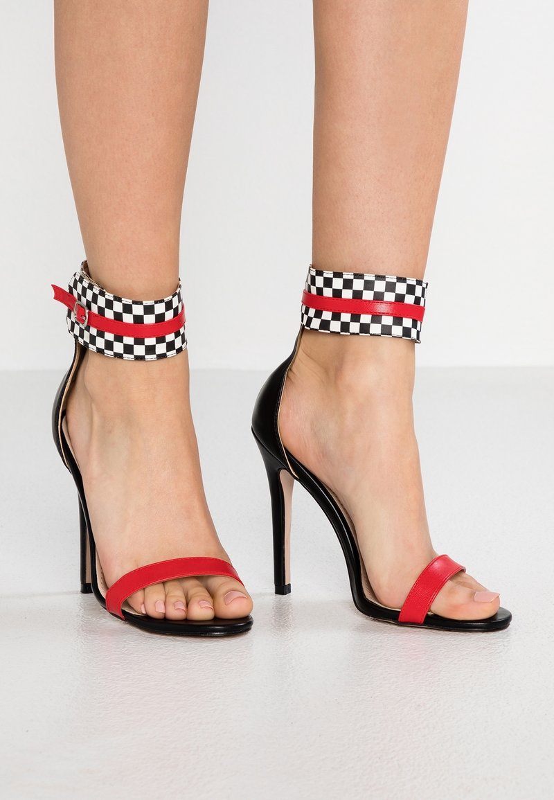 Sandales à talons hauts noires avec des brides rouges sur les orteils et des bracelets de cheville à carreaux noir et blanc ornés de rayures horizontales rouges.