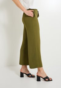 Pantalons larges vert olive en tissu texturé. Dotés d'une taille élastique, de poches et associés à des sandales à talons blonds noires.