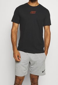 Černé tričko Nike s červeným logem, spárované se světlými šedými sportovními šortkami s černým swoosh znakem Nike. Tkanina vypadá jemně, volný střih.