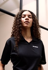Camisa polo preta com um padrão de diamante texturizado. Apresenta colarinho e mangas curtas. Um pequeno logótipo é visível na zona do peito.