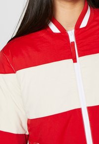 Giacca rossa e crema con zip, caratterizzata da strisce orizzontali. Presenta un colletto a coste e una consistenza simile a un tessuto leggero.