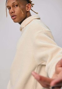 Pull en polaire crème avec un col et de longues manches amples, doté d'une texture douce et de détails de couture minimaux.