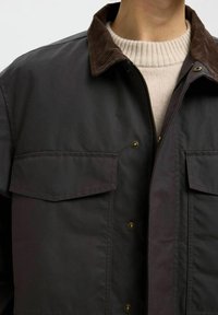 Mörk canvasjacka med en kordflorskrage, som har två bröstfickor och knapp i mässing, lagd över en krämfärgad, ribbad tröja.