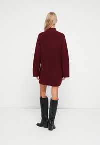 Burgundska pletena obleka z visokim ovratnikom in oversized rokavi, v kombinaciji s črnimi škornji do kolen na peto. Minimalistična podlaga.