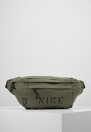 Olive green Nike waist bag with front zipper pocket and adjustable strap, placed on a white pedestal against a plain gray background.