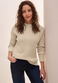 Hellebeige Strickjacke mit einem strukturierten Muster, rundem Halsausschnitt und langen Ärmeln, kombiniert mit dunkelblauen Hosen.