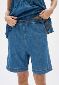 Denim šorti ar elastīgu jostu, brīva piegriezuma un sarullētām piedurknēm. Audums ir vidēji zils ar smalku šuvju detaļām.