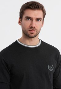 Schwarzes Baumwollhemd mit langen Ärmeln und Rundhalsausschnitt, mit grauem Akzent und kleinem Logo in der oberen linken Ecke.