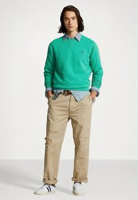 Zöld pulóver, farmer ing, bézs színű, kopott nadrág és fehér, kék részletekkel ellátott sportcipő. A modell zsebbe tett kézzel áll, világos háttér előtt.