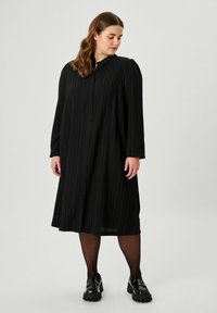 Schwarz, langärmliges Kleid mit vertikalen Falten, knöpfbarem Vorderteil und gewelltem Ausschnitt. Getragen mit schwarzen Strumpfhosen und klobigen Loafers.