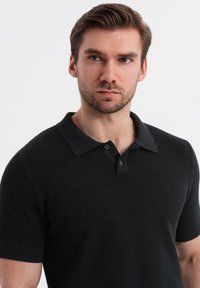 Svart kortärmad pikétröja med en strukturerad stickad mönster, tvåknappsknäppning och klassisk krage. Tyget verkar slätt och åtsittande.