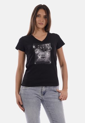 Donna che indossa una maglietta nera a scollo a V con una grafica rettangolare scintillante e jeans grigi, in piedi con una mano nella tasca su sfondo bianco.