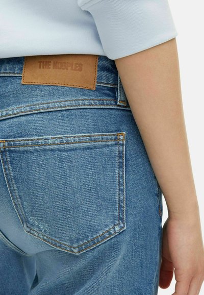 Gros plan sur une personne portant un jean en denim bleu avec une étiquette en cuir « THE KOOPLES » et une chemise à manches longues bleu clair.