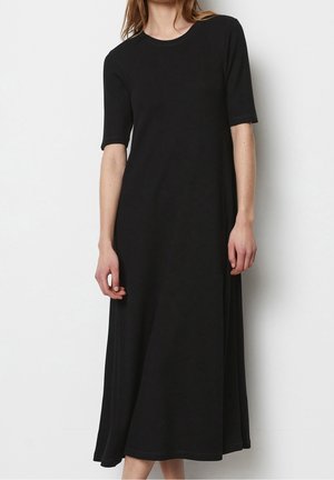 Femme portant une robe midi noire unie à manches courtes, debout devant un fond blanc uni.