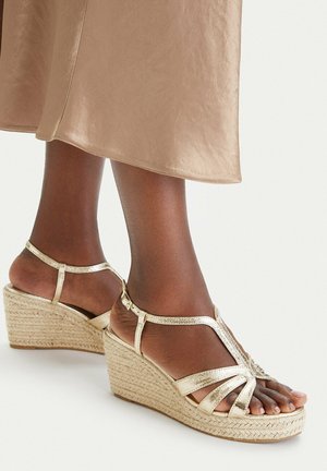 Dunkelhäutige Füße mit beigen Keilsandalen mit Knöchelriemen, kombiniert mit einem glänzenden beigen Midirock.