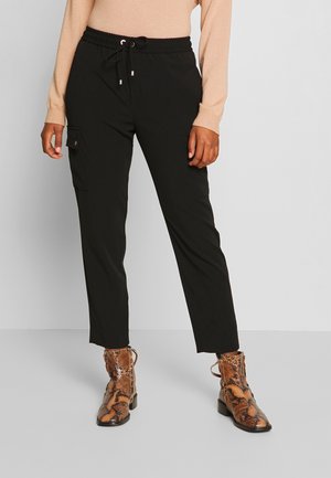 Persoon draagt zwarte cargobroek, beige longsleeve en bruine enkellaarzen van slangenprint, staand op een grijze vloer.