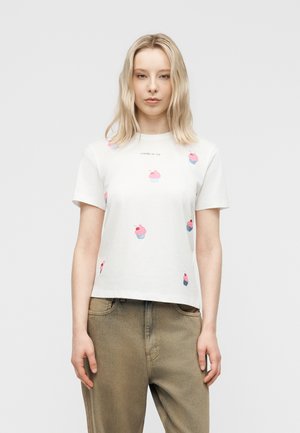 Blondynka w białej koszulce z różowymi nadrukami babeczek i napisem "CHERRY ON TOP", ubrana w luźne beżowe spodnie, stoi na jednolitym tle.