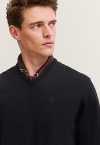 Mann mit kurzen braunen Haaren, der einen schwarzen V-Ausschnitt-Pullover über einem braun gemusterten Schal trägt und nach rechts schaut, vor einem schlichten Hintergrund.