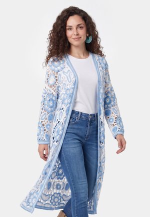 Frau mit lockigem Haar trägt ein weißes Hemd, blaue Jeans und einen langen weißen Cardigan mit blauen Blumenmustern und passenden blauen Ohrringen.