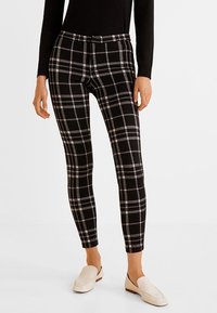 Slim-fit czarne spodnie w białe i szare w kratkę, noszone z czarną bluzką i jasnobeżowymi loafersami, przedstawione od pasa do stóp.